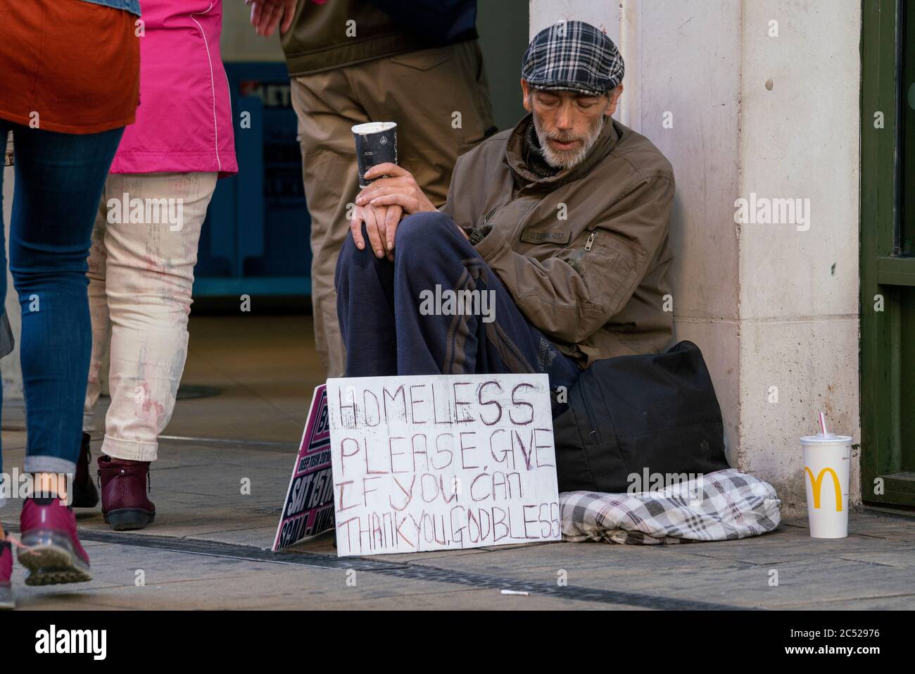 Un uomo senza casa, siede fuori, con un caffè McDonalds nelle sue mani. Con un cartello che chiede denaro. Come la gente cammina, senza preoccuparsi. Foto Stock