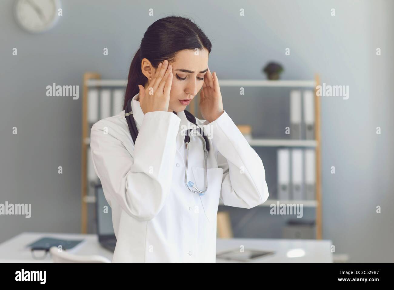 Giovane medico femminile che soffre di mal di testa, affaticamento, pressione alta o privazione del sonno in clinica Foto Stock