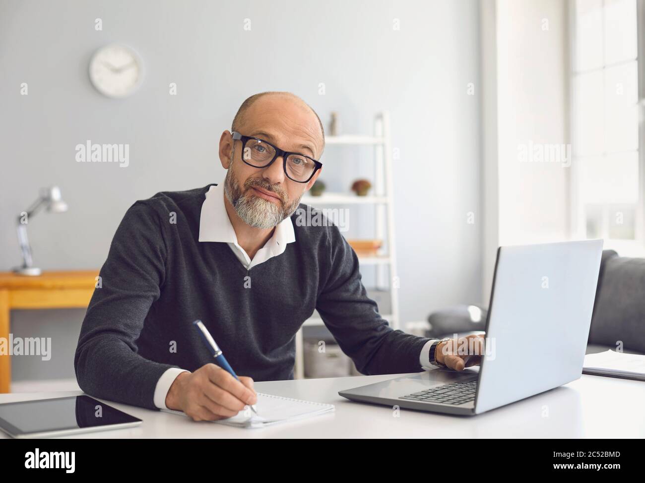 Un uomo anziano con gli occhiali sta lavorando sta studiando ha un laptop che scrive le note in un taccuino mentre siede ad un tavolo nel suo ufficio domestico. Foto Stock