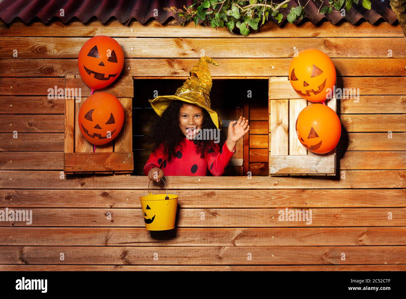 Carino ritratto ragazza in cappello Halloween e costume con il secchio giallo guardare dalla finestra della casa sull'albero Foto Stock