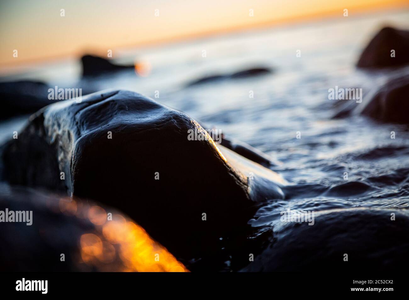 Foto closeup delle onde marine che frena su una riva bagnata pietre estate tramonto, spruzzi d'acqua, raggi solari, riflessi sulle gocce d'acqua. Foto Stock