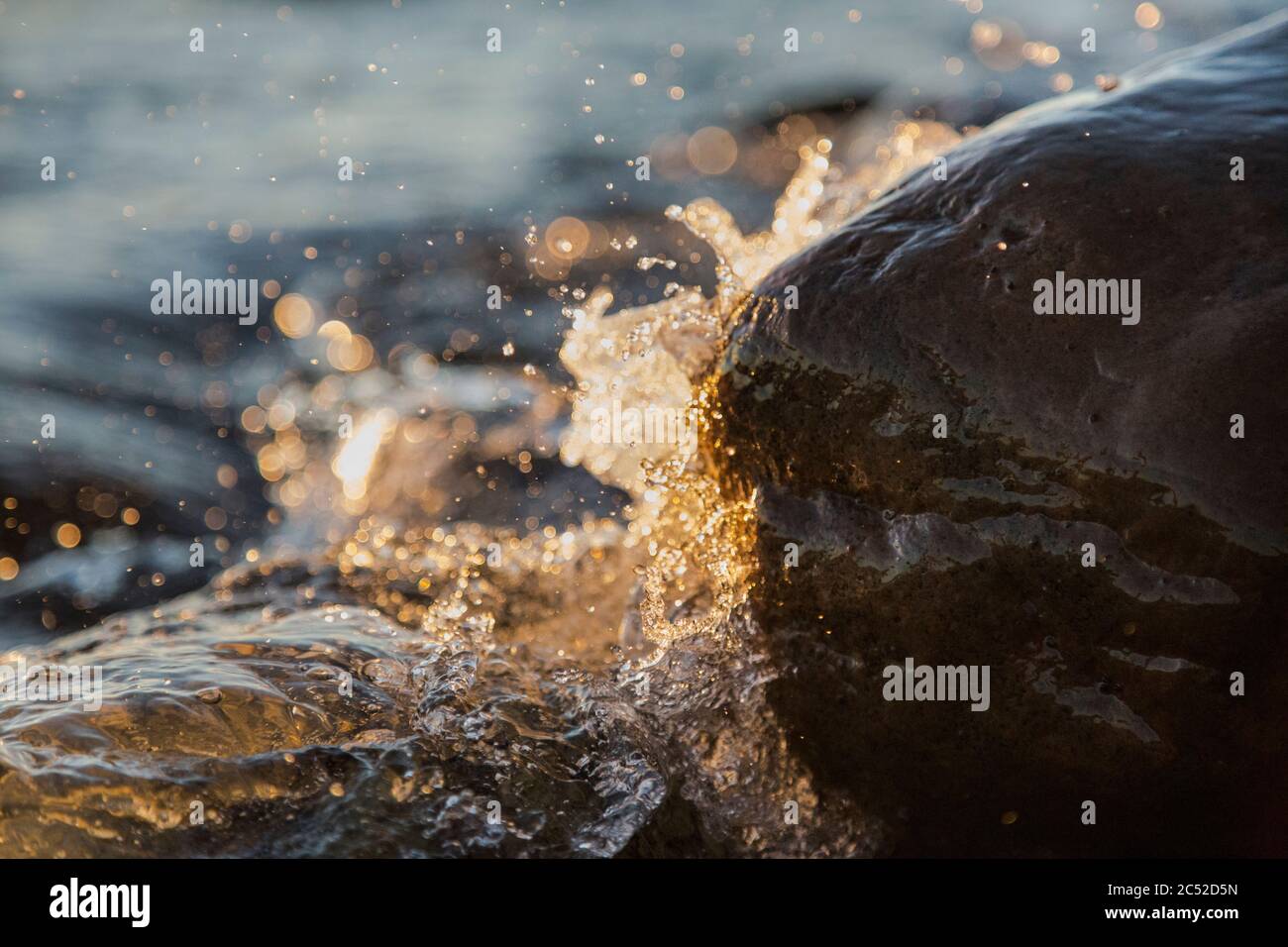 Foto closeup delle onde marine che frena su una riva bagnata pietre estate tramonto, spruzzi d'acqua, raggi solari, riflessi sulle gocce d'acqua. Foto Stock