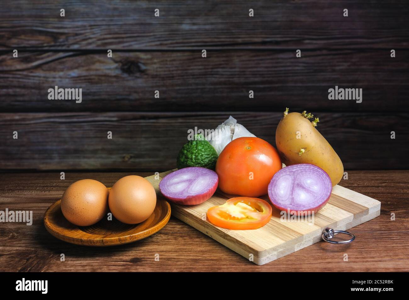 Pomodoro, aglio, lime kaffir, cipolla, uova e patate su un tagliere di legno Foto Stock