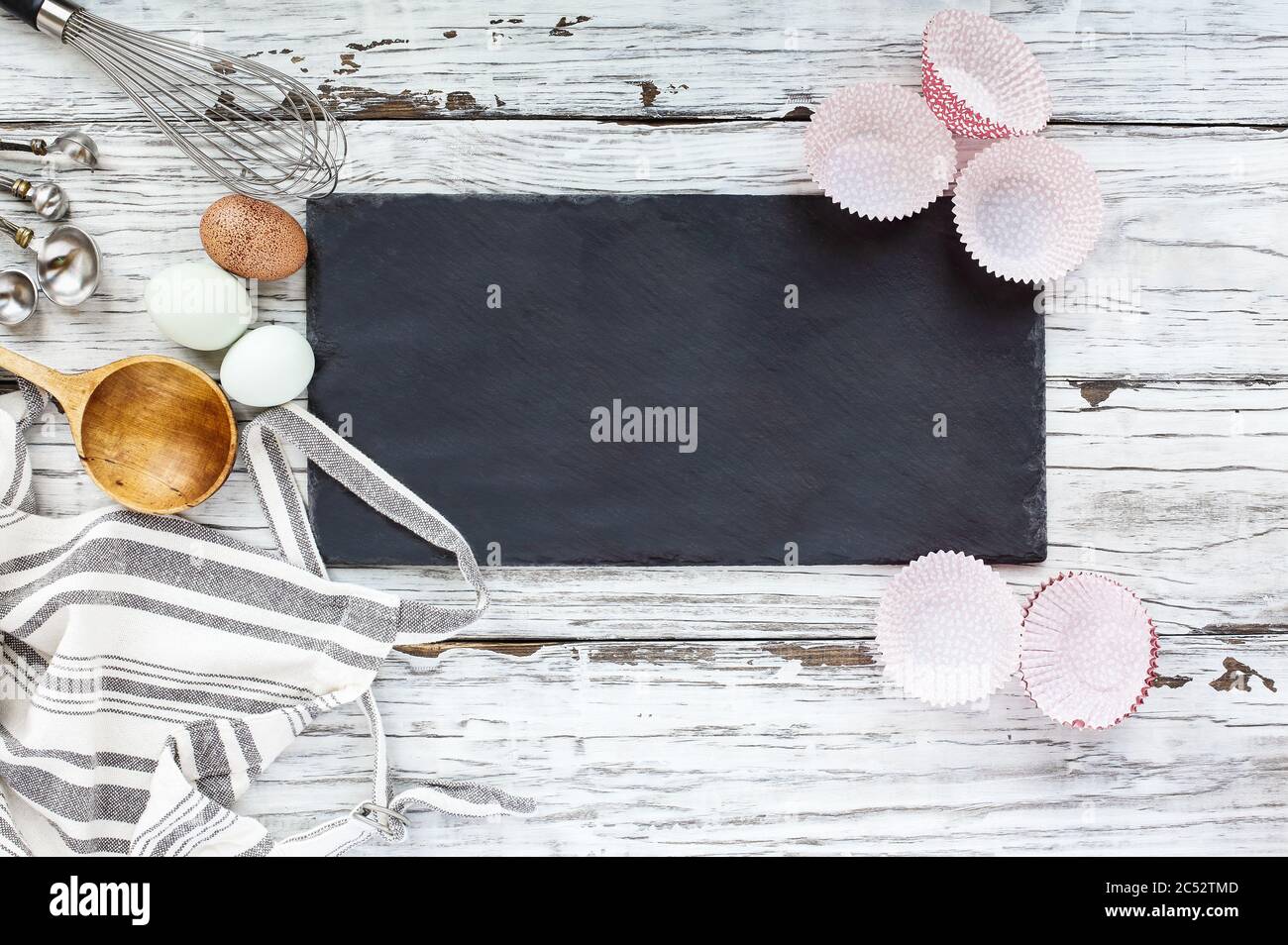 Cartello in bianco con fodere, grembiule, frusta, misurini, cucchiaio vecchio di legno e uova su sfondo bianco di legno. Immagine ripresa dalla vista superiore Foto Stock