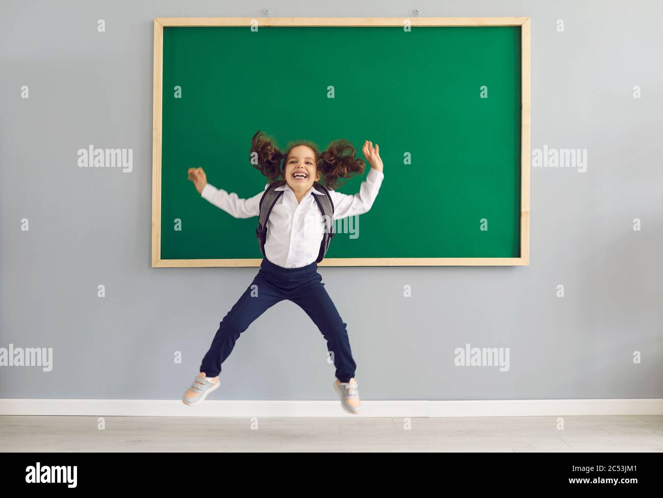 Concept prima volta a scuola. Ragazza divertente con uno zaino sta saltando sullo sfondo della scuola in classe. Foto Stock