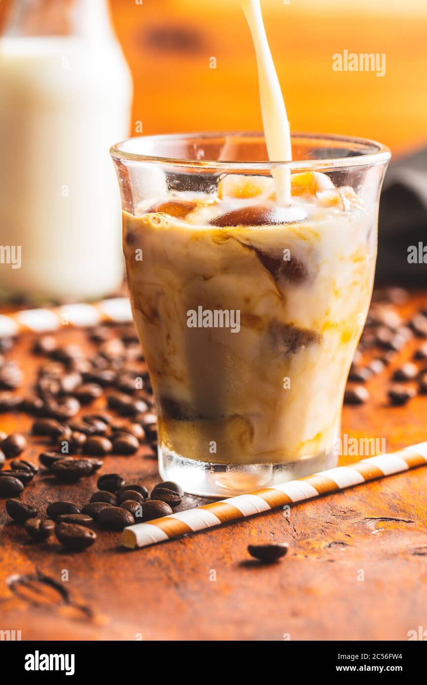 Versare il latte in un bicchiere con caffè ghiacciato su un tavolo di legno. Foto Stock