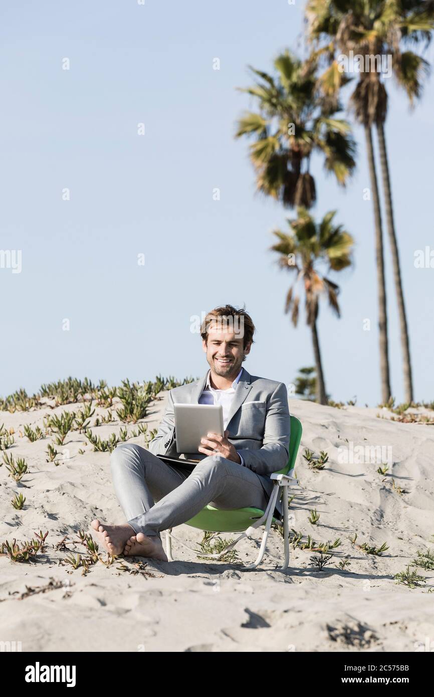 Uomo d'affari a piedi nudi che usa un tablet digitale sulla spiaggia soleggiata, Los Angeles, California Foto Stock