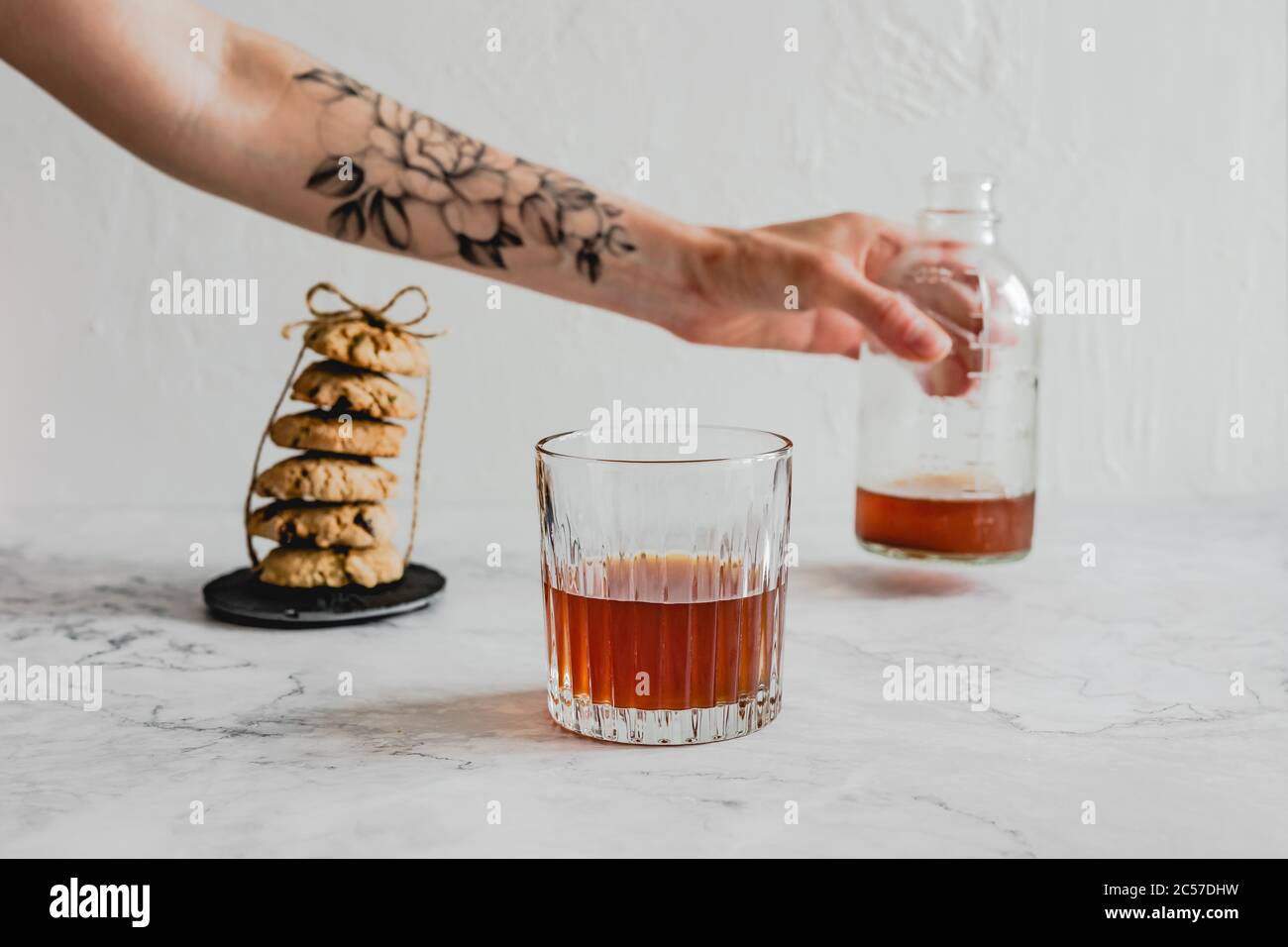 Bielorussia, Minsk - 19.06.2020: Un bicchiere di caffè freddo, una pila di biscotti d'avena e una mano con tatuaggio floreale che contiene una bottiglia Foto Stock