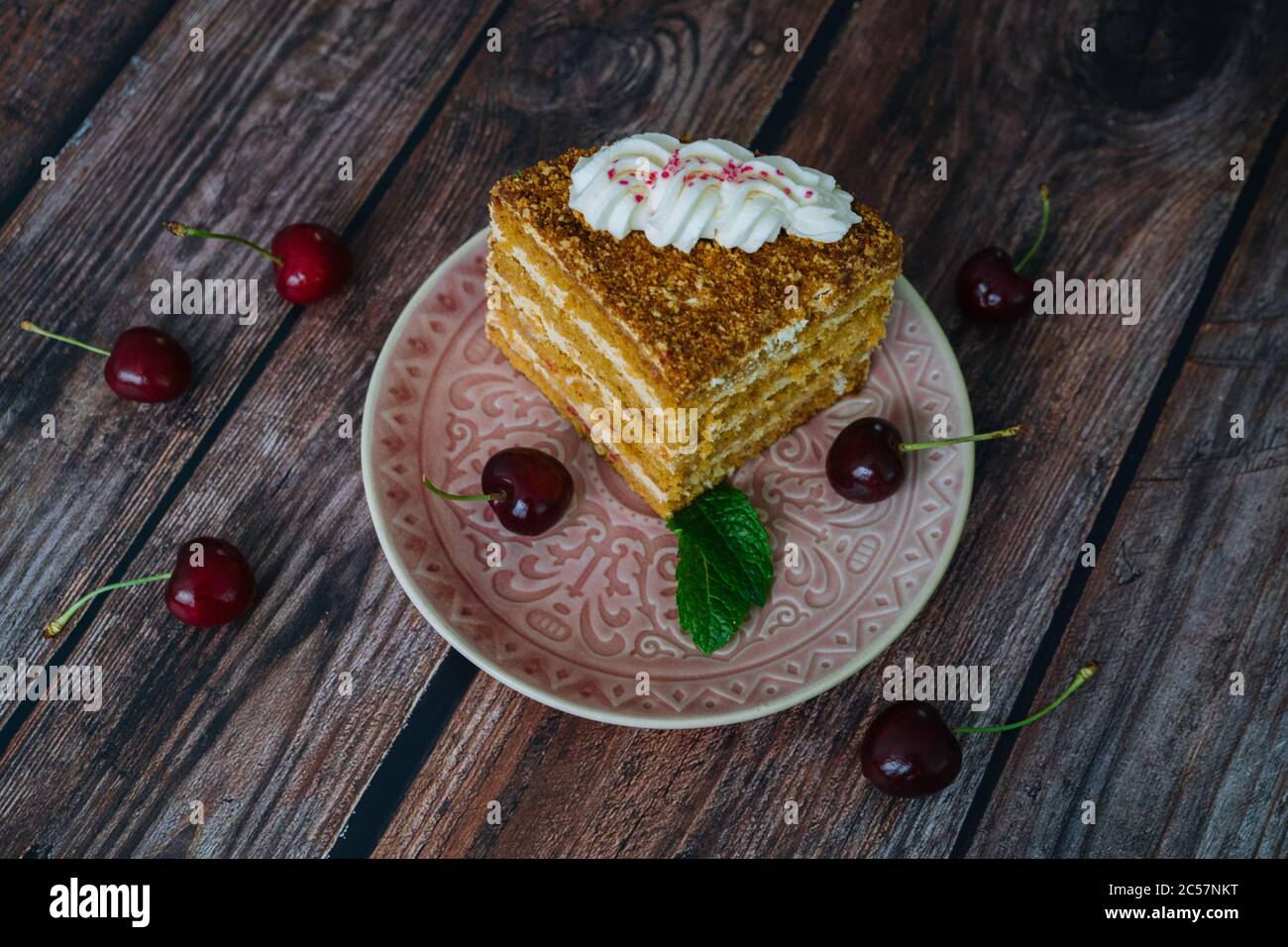 Pezzo di torta al miele multistrato con crema. Classici pezzi di torta al miele con crema pasticcera. Torta al miele fatta in casa su sfondo di legno con foglie di ciliegia e menta. Deliziosa fetta di torta a strati sul piatto Foto Stock