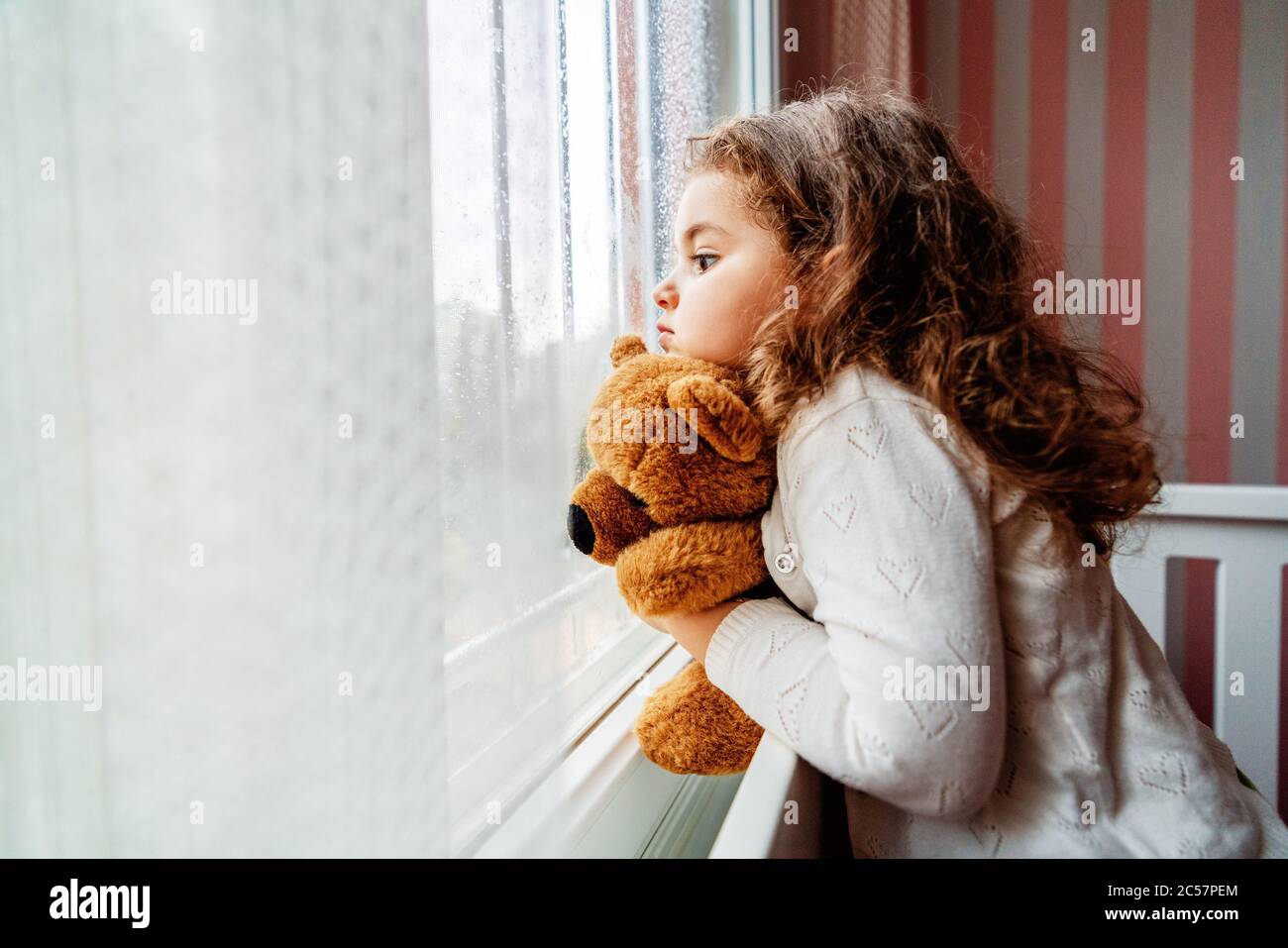 La ragazza carina piccola sta guardando dalla finestra alla pioggia con tristezza si annoia. . Foto di alta qualità Foto Stock