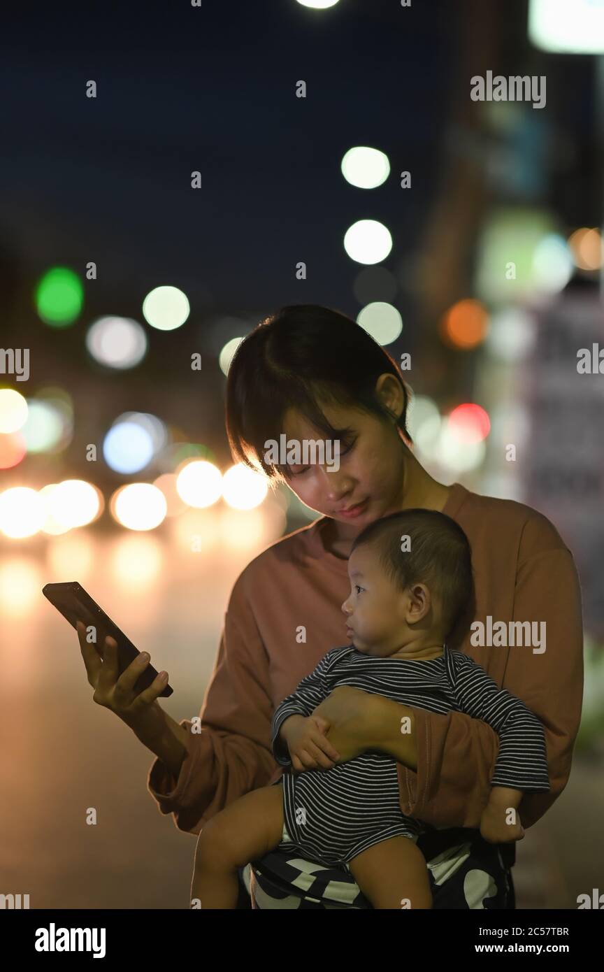 Donna e bambino asiatici aspettano il suo taxi privato utilizzando un'app di trasporto sulla strada notturna. Foto Stock