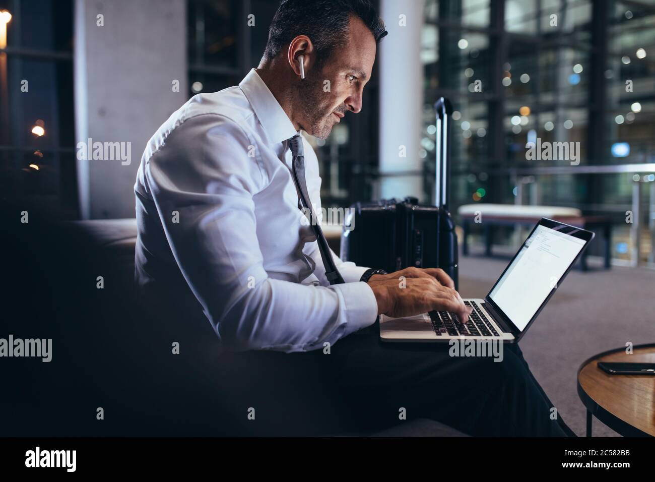 Uomo d'affari che lavora su un computer portatile mentre attende il suo volo in aeroporto. Uomo in abbigliamento da lavoro che utilizza un computer portatile nell'area d'attesa dell'aeroporto. Foto Stock