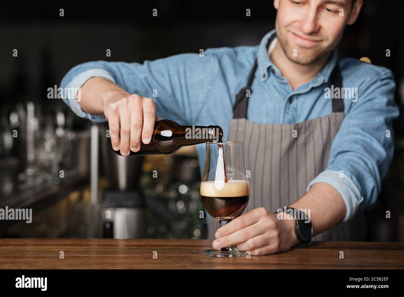 Ripetere l'ordine. Barman in grembiule e camicia in denim, versa la birra scura in vetro Foto Stock