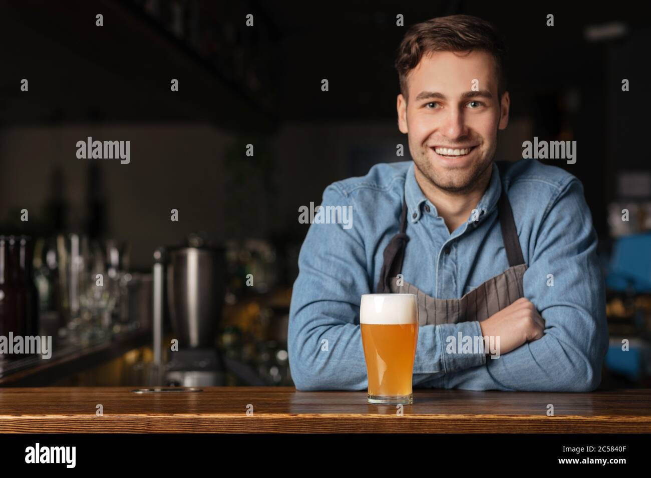 Tradizione della birra in un pub moderno. Un bel ragazzo sorridente in camicia in denim si appoggia al bancone bar con vetro Foto Stock