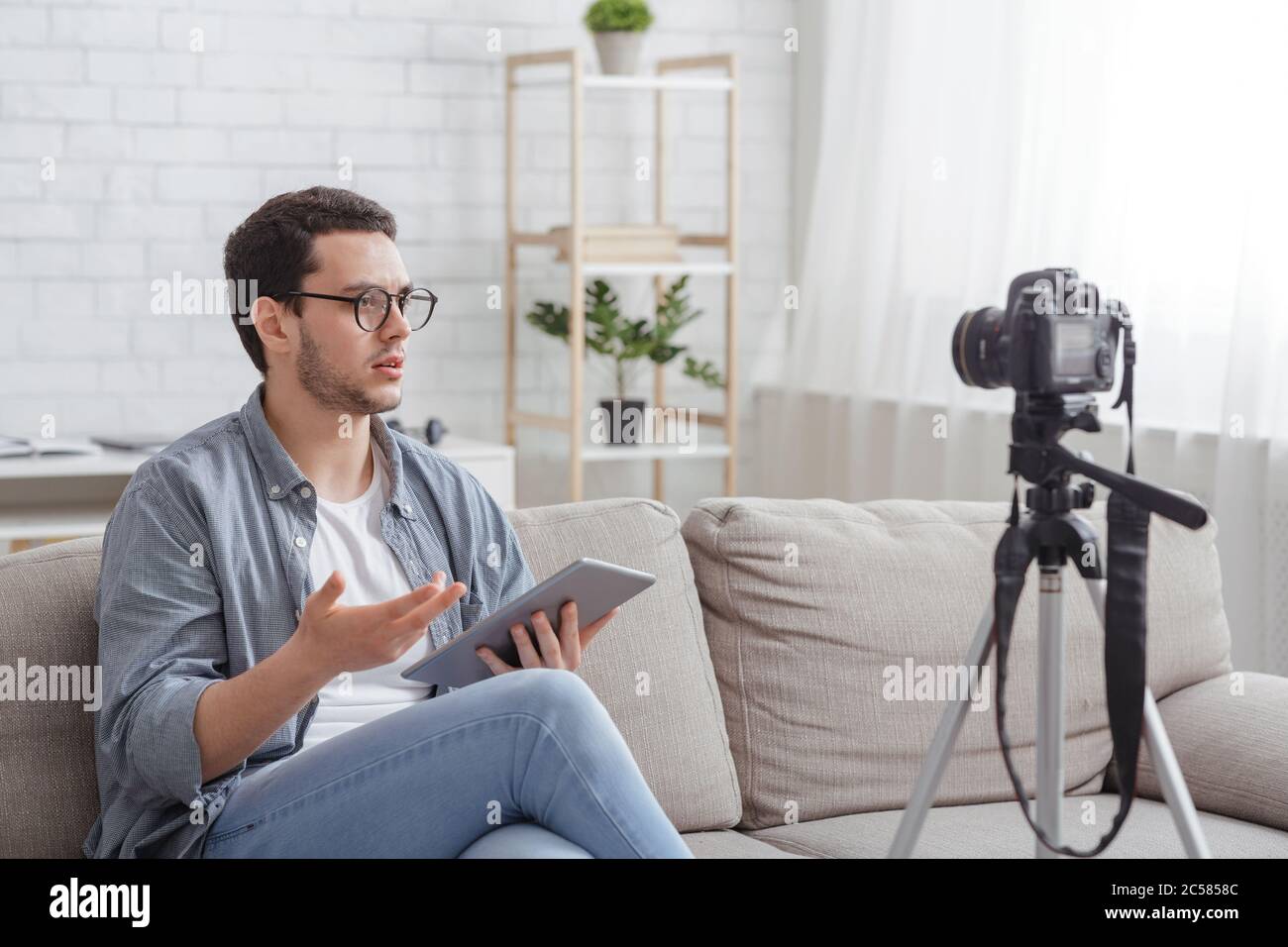 Blogger opinione. Un uomo serio con il tablet in mano si siede sul divano, fa video Foto Stock