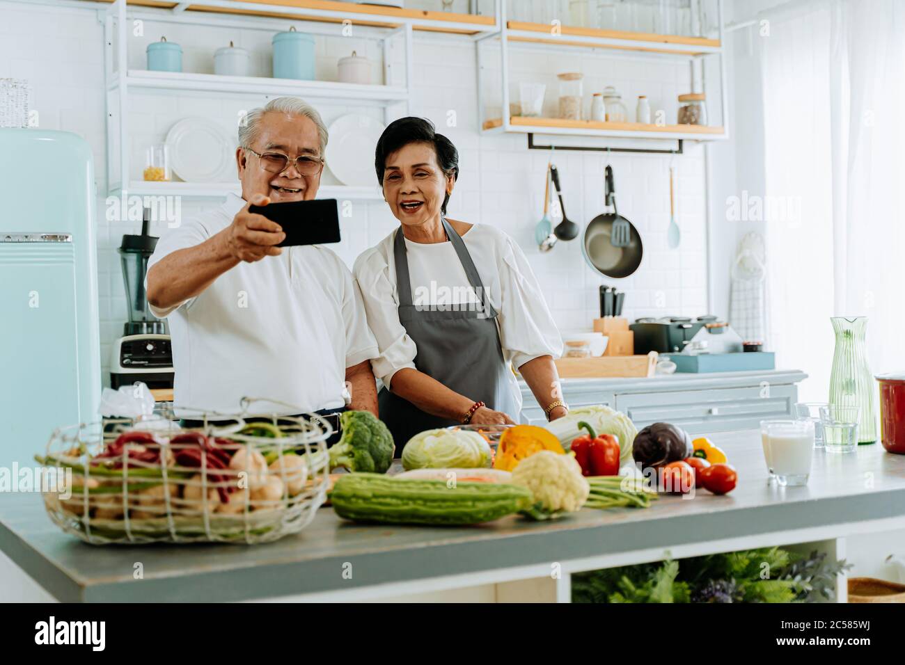 Allegro anziano uomo e donna asiatici sorridendo e salutando amico mentre si effettua una videochiamata su smartphone e cucinare in cucina Foto Stock