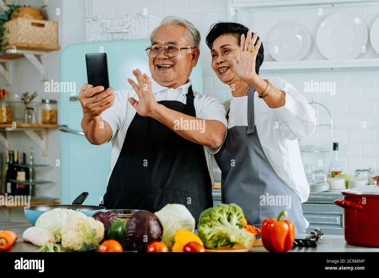 Allegro anziano uomo e donna asiatici sorridendo e salutando amico mentre si effettua una videochiamata su smartphone e cucinare in cucina Foto Stock