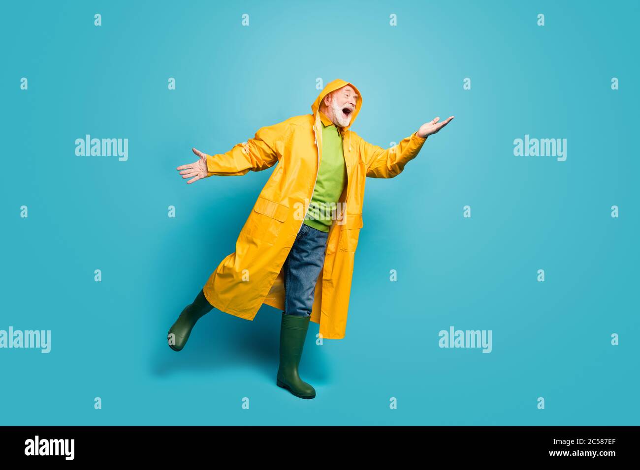 Vista a lunghezza intera del corpo del suo bel bel ragazzo positivo grigio-capelli che indossa il topcoat giallo cattura goccia piovosa avendo divertimento isolato su luminoso Foto Stock