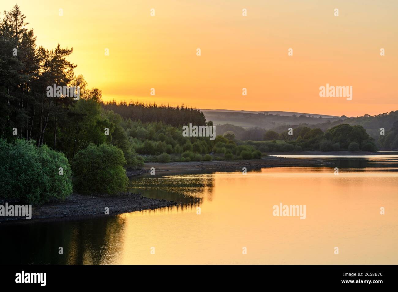 Splendido tramonto panoramico rurale su acque tranquille (alberi boschivi e riflessi di colorato cielo arancione) - Fewston Reservoir, North Yorkshire, GB. Foto Stock