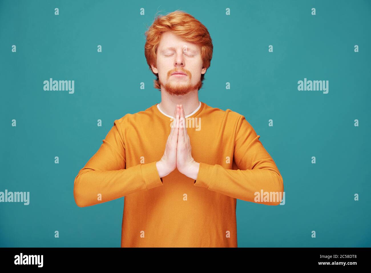 Calma spirituale giovane uomo rosso con barba facendo gesto Namaste meditando con occhi chiusi su sfondo blu Foto Stock