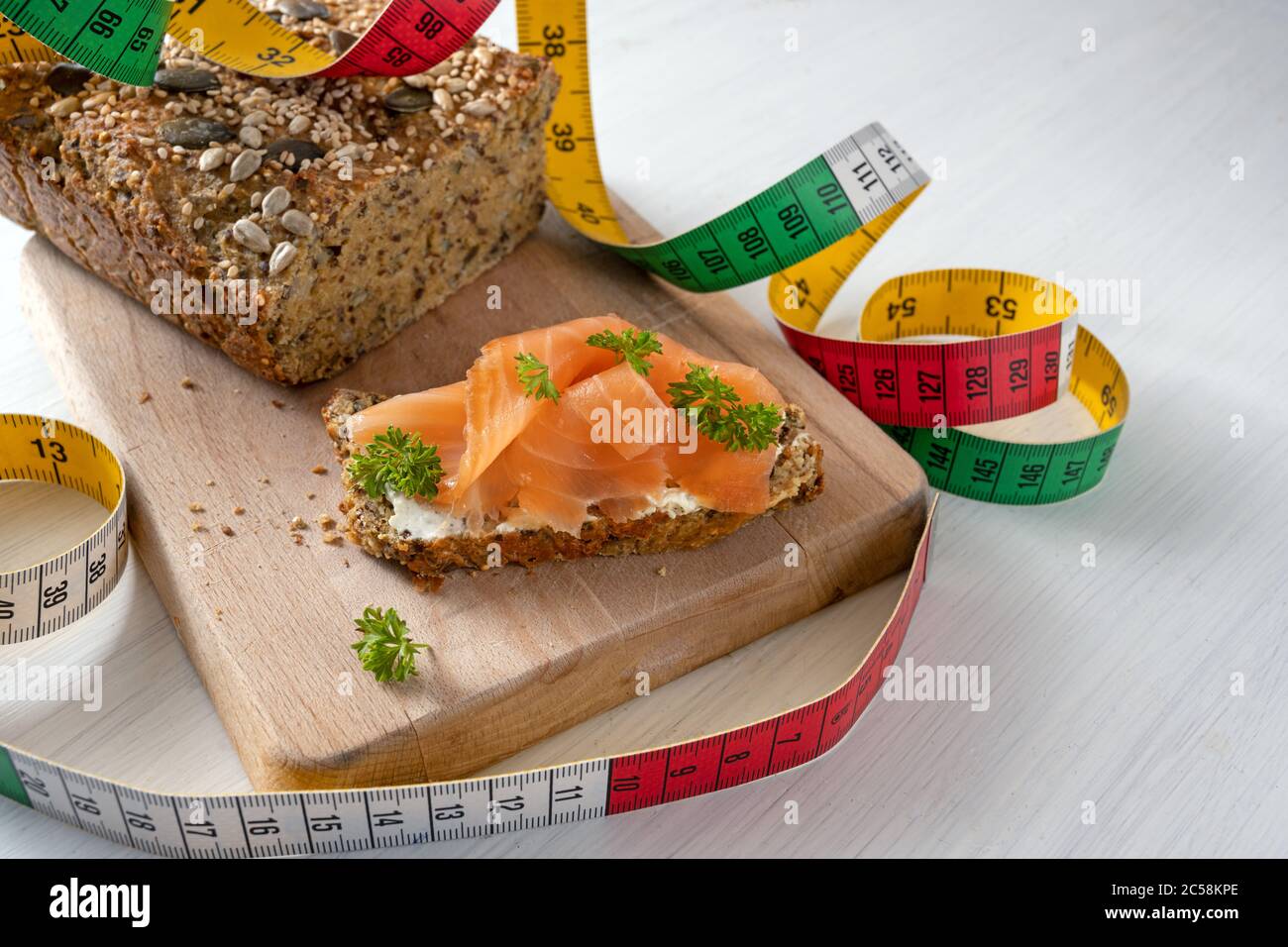 Dimagrire con dieta a basso contenuto di carb o chetogenico, pane proteico con metro a nastro e un sandwich con guarnitura di salmone e erbe su un tagliere e su un vortico Foto Stock