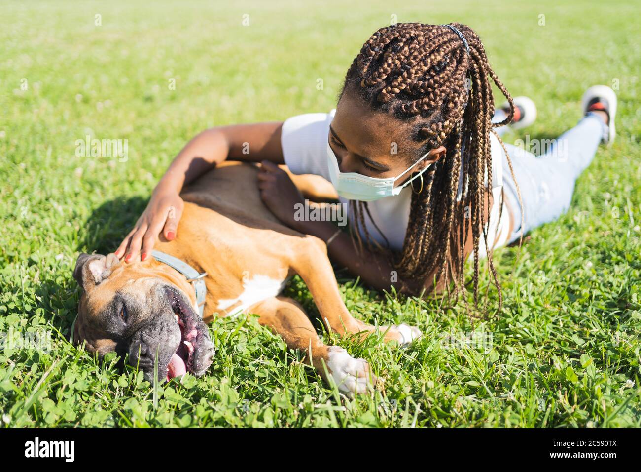 Donna africana con trecce e maschera protettiva stesa sull'erba e stropica un cane pugile in una giornata di sole Foto Stock