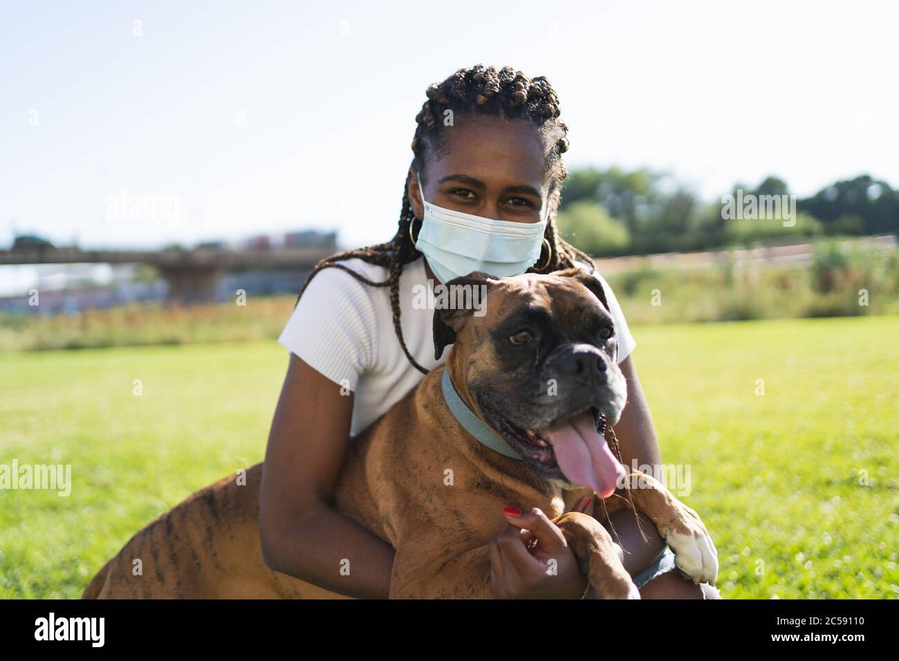 Donna africana con trecce e maschera protettiva che abbracciano un cane da boxe con le braccia in erba in una giornata di sole Foto Stock