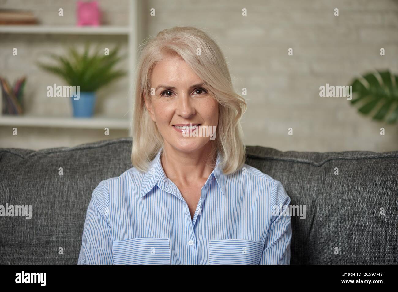 Bella donna bionda di mezza età con un sorriso luminoso seduto su un divano a casa Foto Stock