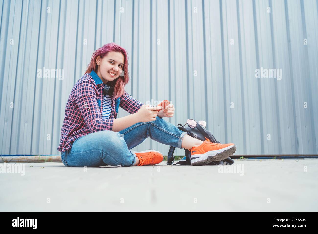 Bella giovane adolescente moderna sorridente in una camicia a scacchi e jeans con cuffie e smartphone seduto sul marciapiede di strada. Moderno Foto Stock