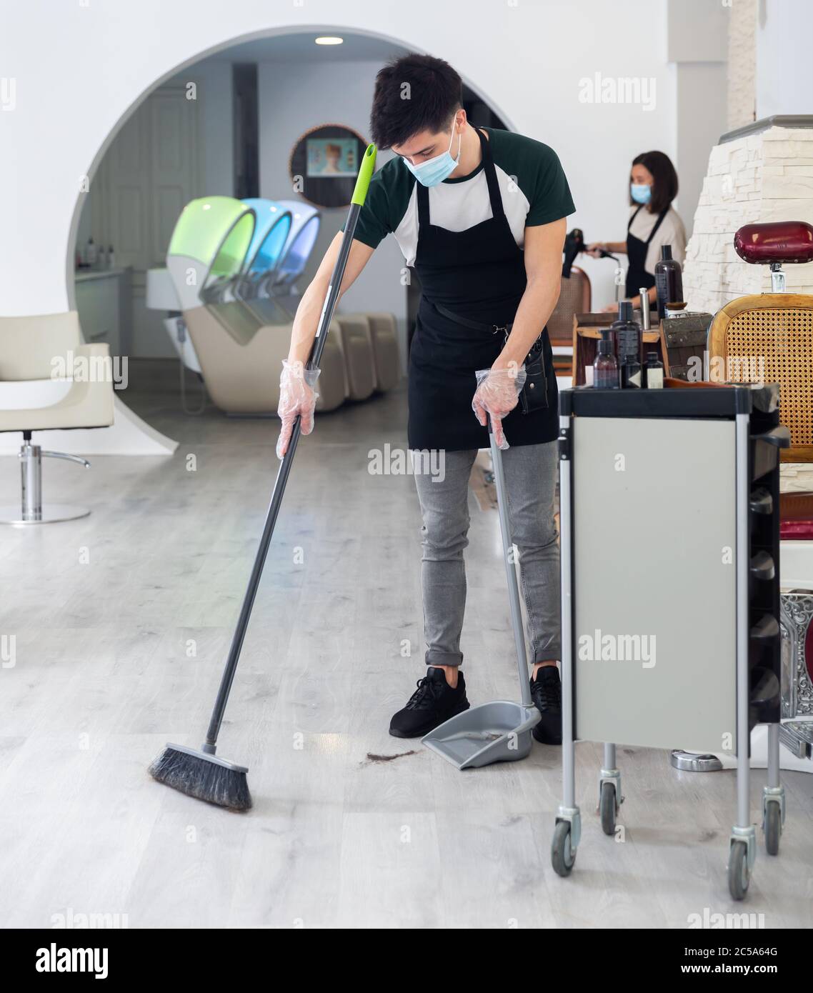 Processo di lavoro in studio di bellezza durante la situazione pandemica, uomo spazzare pavimento, donna hairstyler fare capelli al cliente sullo sfondo, tutte le persone che Foto Stock