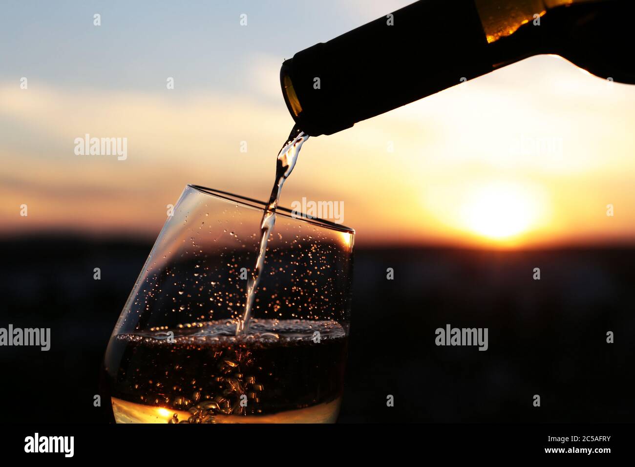 Vino bianco che si versa da una bottiglia nel bicchiere su uno sfondo bellissimo tramonto, fuoco selettivo. Concetto di celebrazione, cena romantica Foto Stock