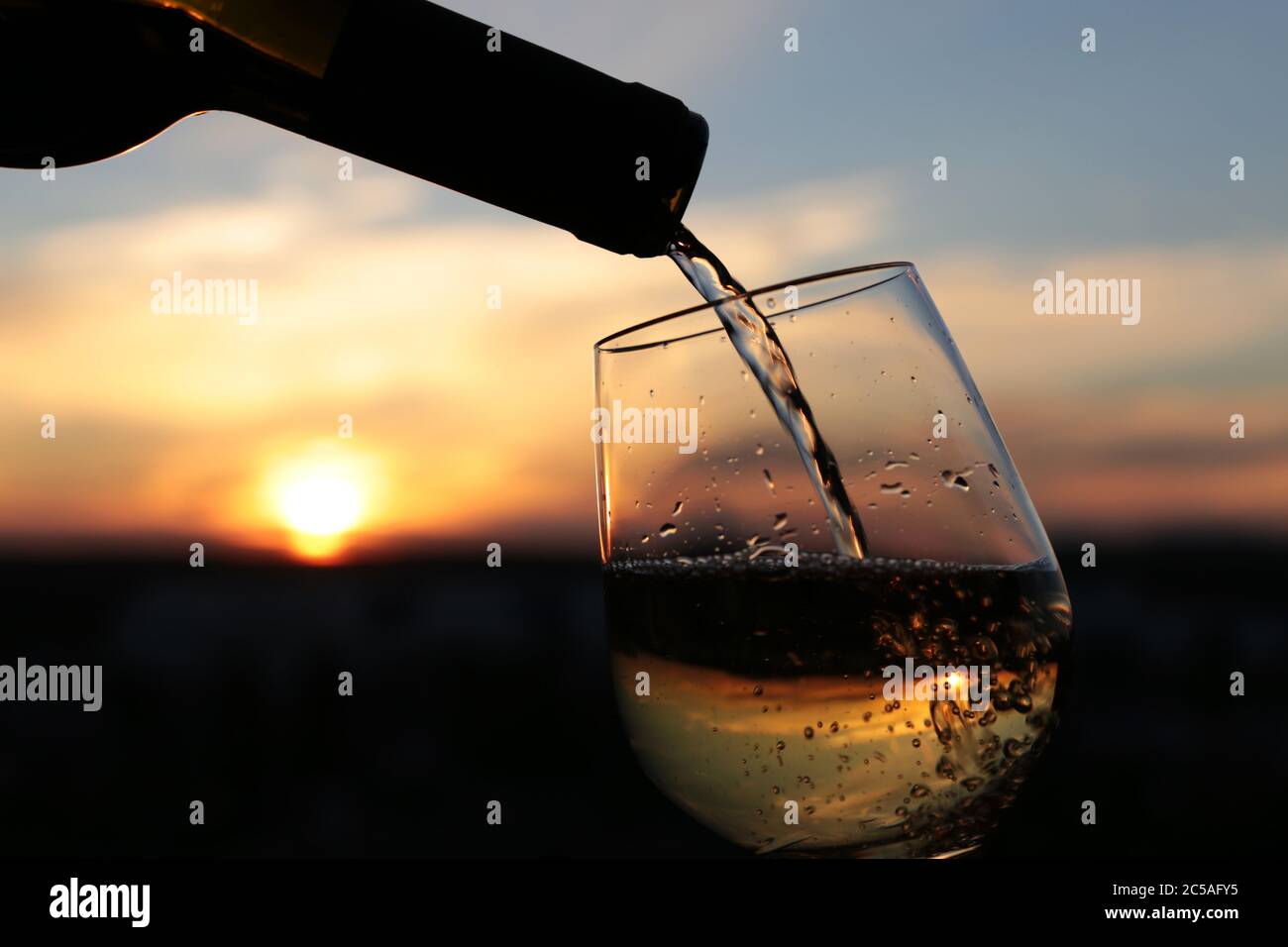 Vino bianco che si versa da una bottiglia nel bicchiere su uno sfondo bellissimo tramonto, fuoco selettivo. Concetto di celebrazione, cena romantica Foto Stock