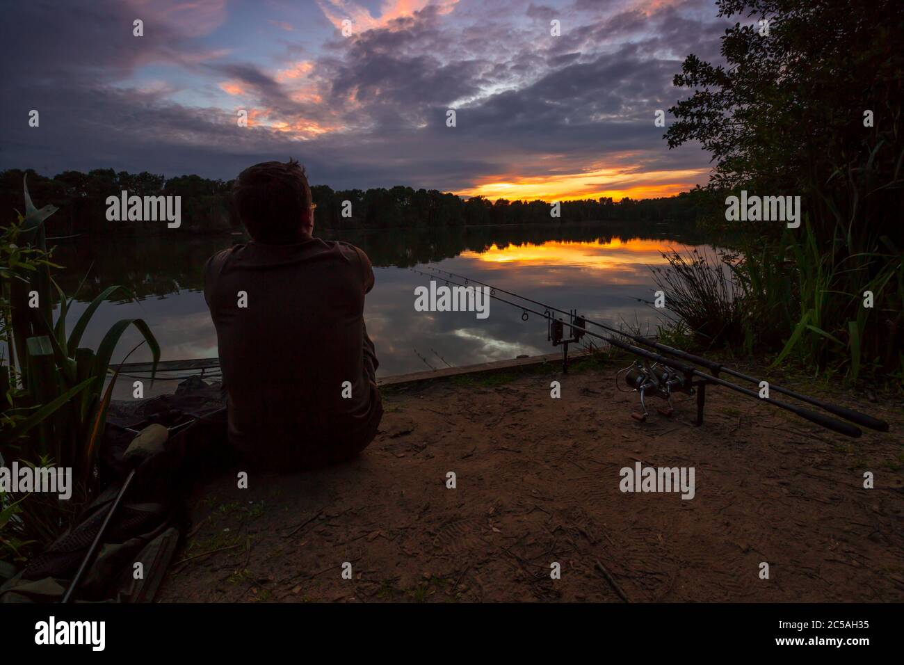 Uomo carpa pesca su un lago guardando il tramonto seduto accanto ad alcune canne da pesca Foto Stock