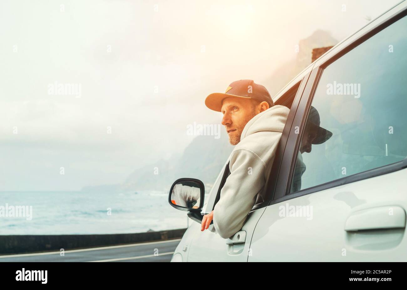 Uomo vestito caldo felpa con cappuccio e cappello da baseball che guida la sua nuova auto sulla strada solitaria che conduce dalla costa del mare. Immagine di concetto del proprietario dell'auto felice. Foto Stock