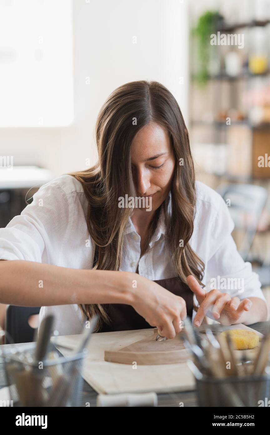 Donna che lavora con l'argilla con le mani e l'utensile da taglio ad anello da vicino in un laboratorio di ceramica. Foto Stock