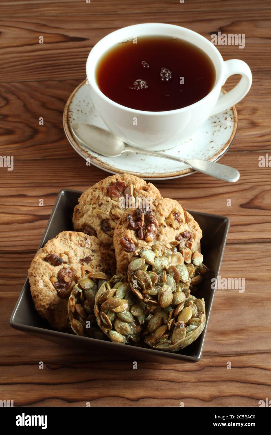 Biscotti di farinata d'avena con noci e uvetta, biscotti di semi di zucca ricoperti di zucchero in una ciotola quadrata e una tazza di tè nero su un tavolo di legno. Primo piano Foto Stock