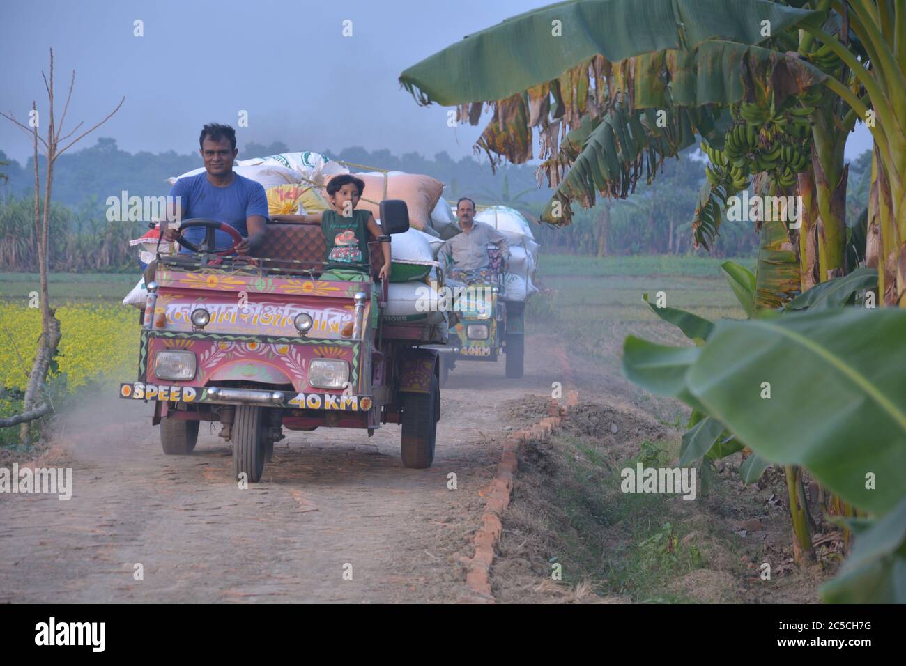 Un uomo che guida un furgone motore o un risciò motorizzato con un passeggero e merci in una strada di villaggio, focalizzazione selettiva Foto Stock