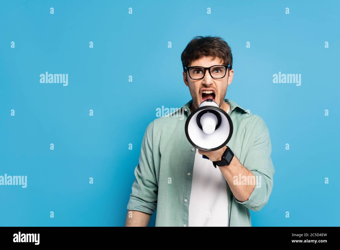 giovane arrabbiato urlando in megafono mentre si guarda la telecamera su blu Foto Stock
