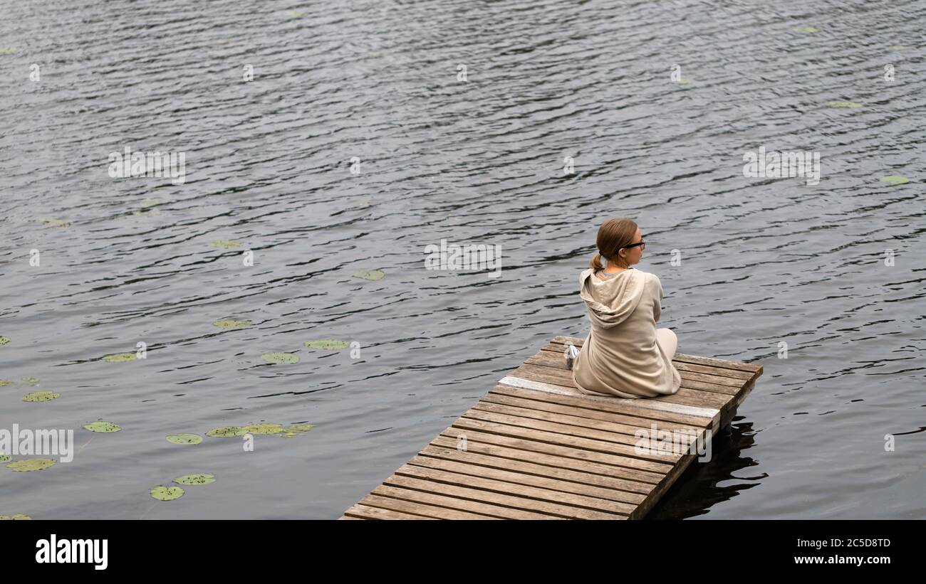 Giovane donna seduta sul molo di legno, riposante e guardando il fiume, vista dall'alto. Ora legale. La gioia della vita di campagna. Foto Stock