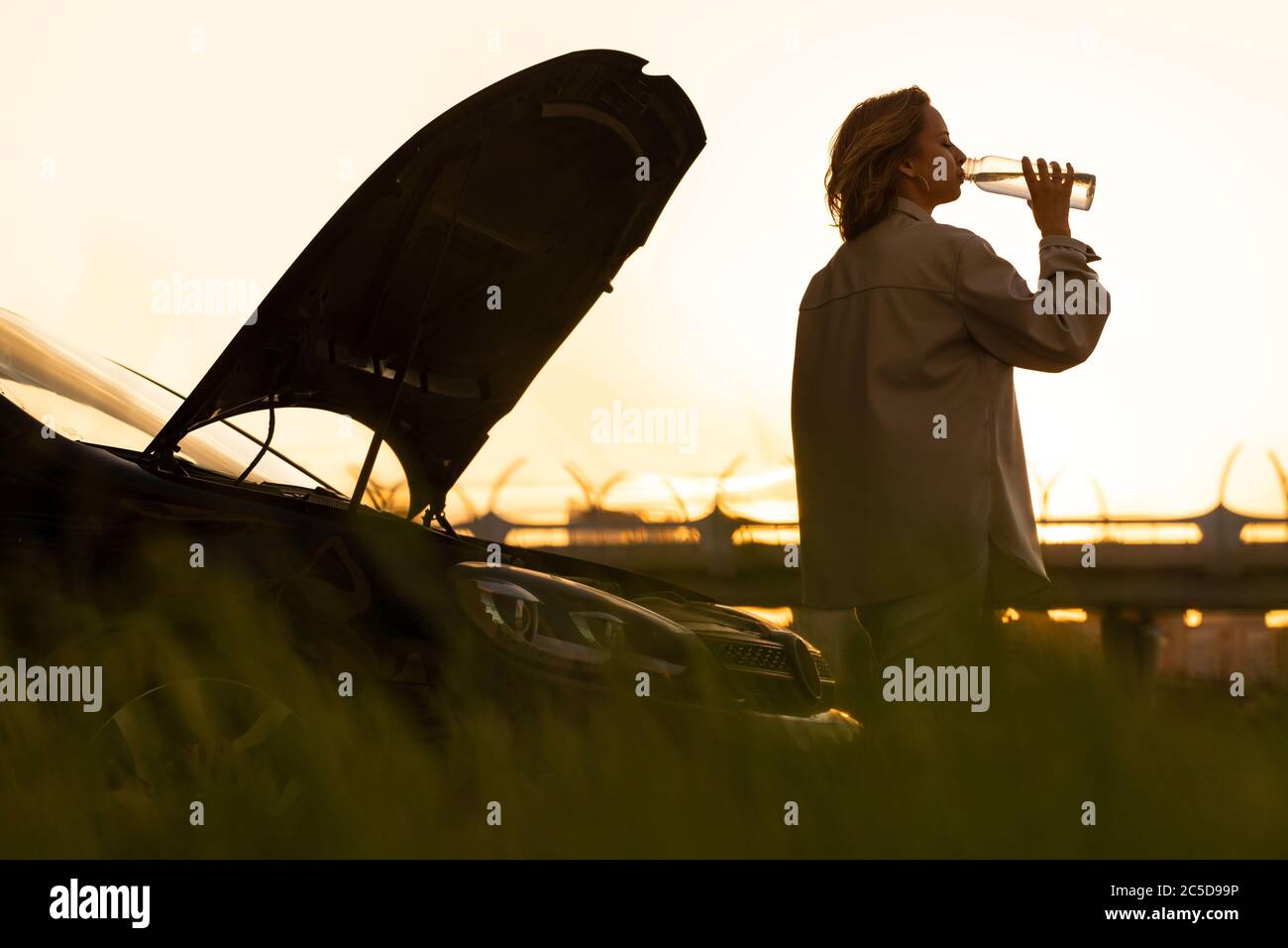 Silhouette di donna che beve acqua da una bottiglia ricaricabile, stanco di aspettare l'assistenza vicino alla sua auto rotto con cofano aperto al tramonto. Foto Stock