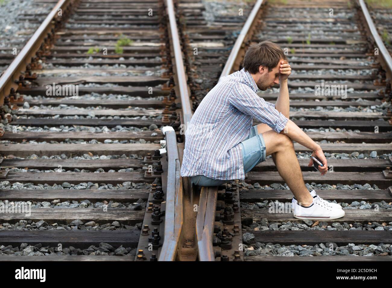 Uomo depresso seduto sulle piste ferroviarie, tenendo il telefono, prende una decisione difficile di vivere nel passato o cambiare il suo futuro. Cambiare il percorso di vita c Foto Stock