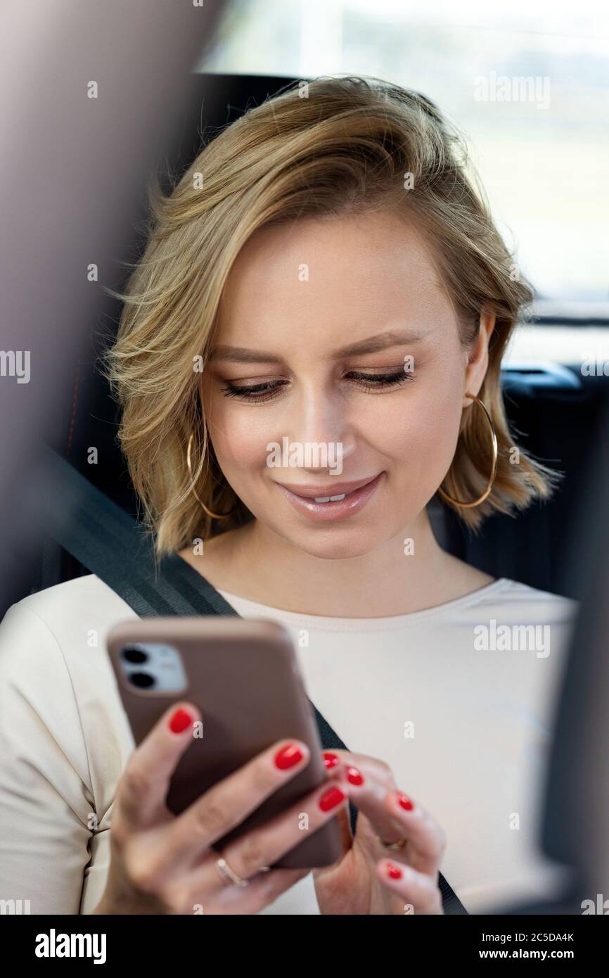 Donna sorridente seduto in auto sul sedile passeggero allacciato da una cintura di sicurezza, utilizzando smartphone, scorrimento dei social network, lettura dei messaggi. Primo piano. Foto Stock