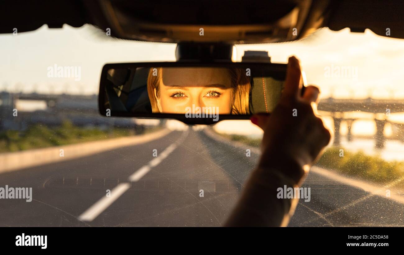 Donna allegra e gioiosa che regola lo specchio mentre si siede in auto, guardando in riflessione la fotocamera. Ora del tramonto. Emozioni dalla guida dell'auto Foto Stock
