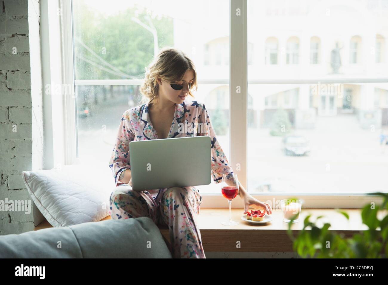 Bere vino. Ritratto di giovane ragazza in appartamento moderno al mattino. Calma, salificata. Concetto di gioventù e benessere. Donna elegante in pajama con occhiali che si adora comodamente con il laptop. Foto Stock