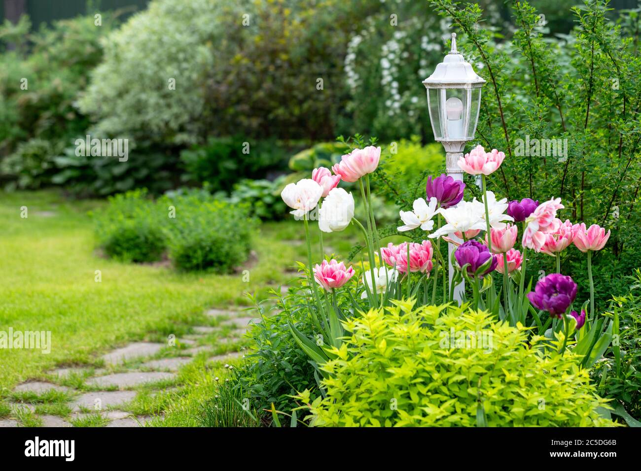 Lampada da giardino decorativa d'epoca circondata da tulipani in fiore, erba, cespugli, passerella in pietra. Cottage giardino Foto Stock