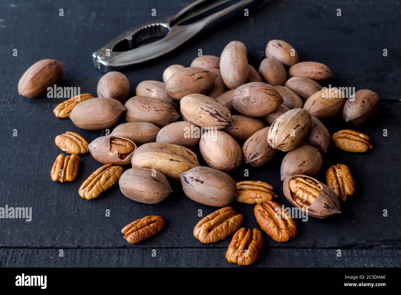 Noci di pecan in guscio, spaccate e metà isolate su rustico sfondo di legno nero con nutcracker de-focalizzato sullo sfondo Foto Stock