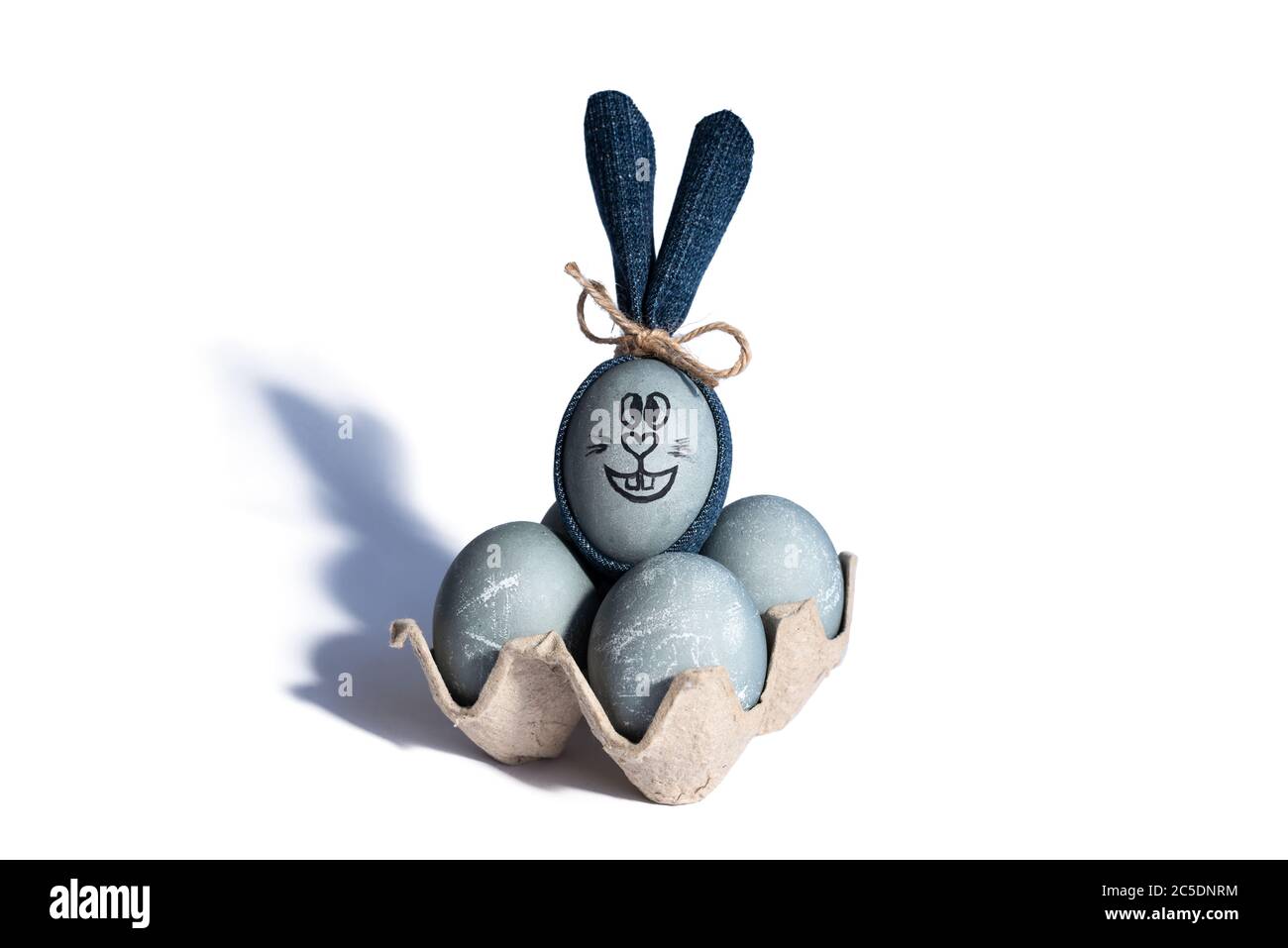 Uova di Pasqua a colori di cemento in vassoio di cartone e coniglietto pasquale con orecchie di denim blu su sfondo bianco. Buon concetto di Pasqua. Idea fai da te Foto Stock