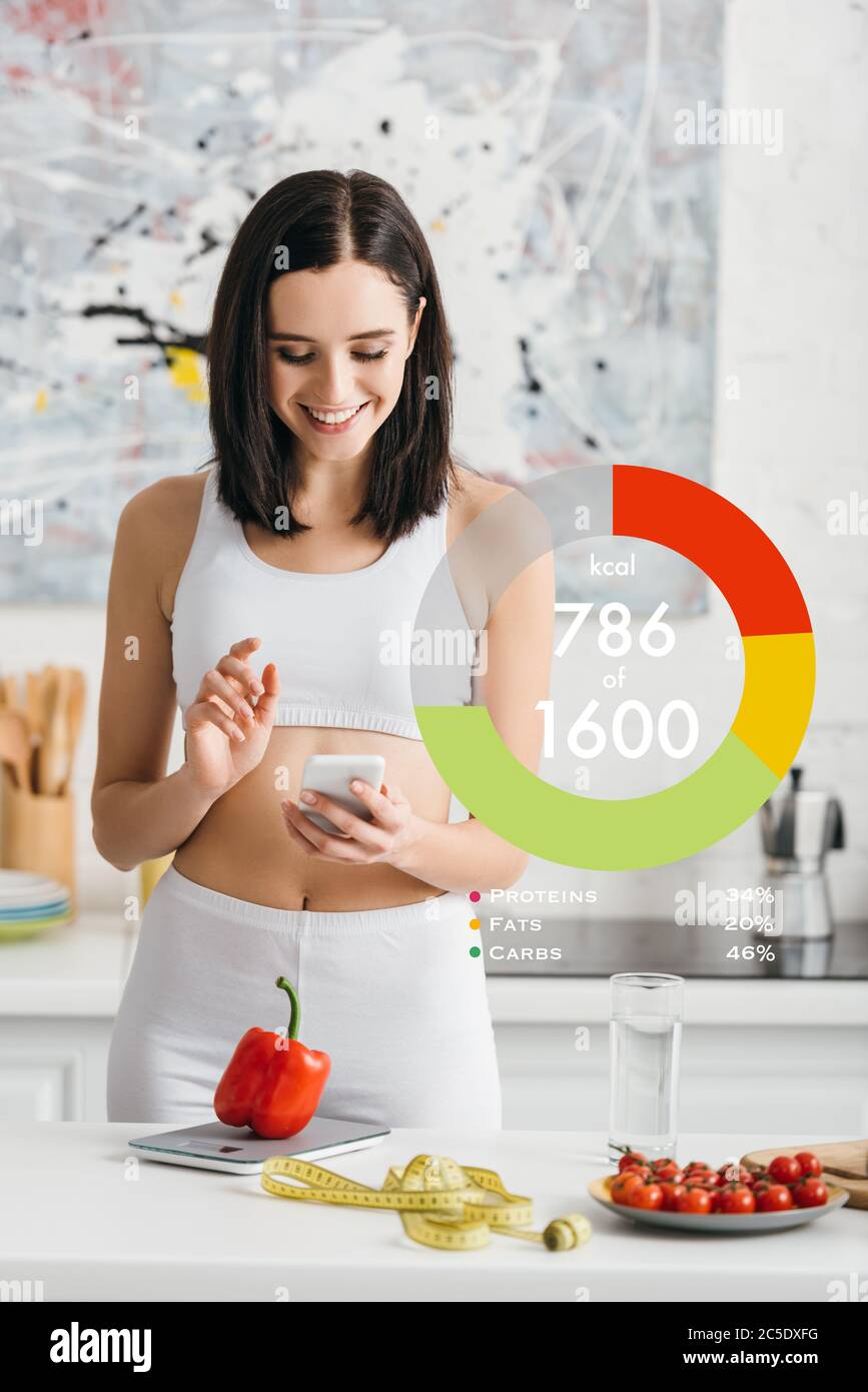 Donna sportiva sorridente che usa uno smartphone vicino a nastro di misurazione, verdure e bilance su tavolo da cucina, illustrazione del conteggio delle calorie Foto Stock