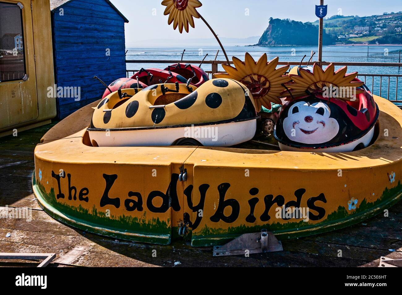 Le carrozze a due posti dipinte di rosso e giallo di un giro per bambini in Ladybirds si siedono in modo forgnoso sul loro percorso sul molo di Teignmouth con il mare blu alle spalle Foto Stock