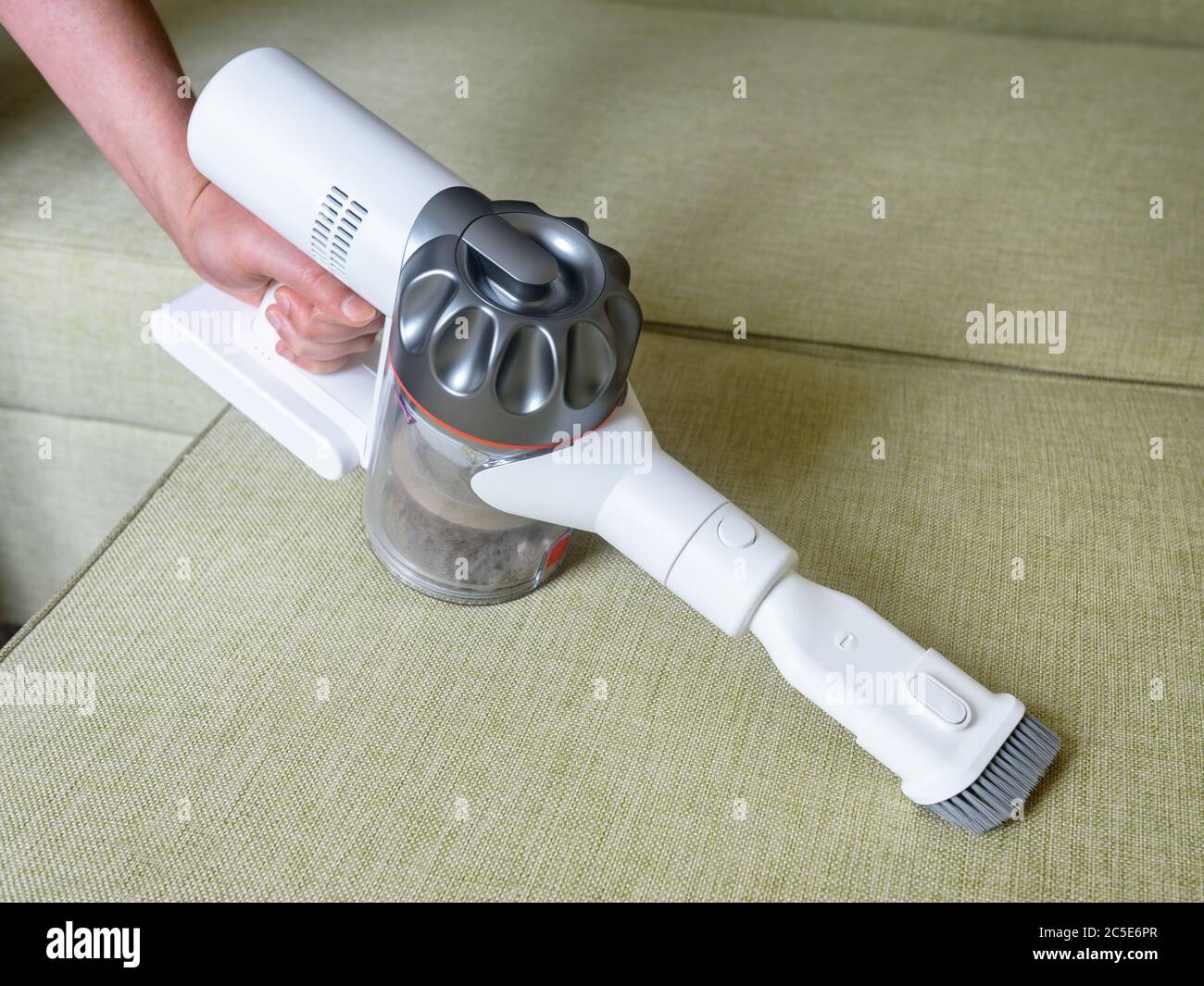 Aspirapolvere wireless utilizzato sul divano in camera. Lavori domestici con nuovo gancio verticale e bocchetta. La persona può utilizzare un aspirapolvere bianco moderno. Pulizia della casa, c Foto Stock